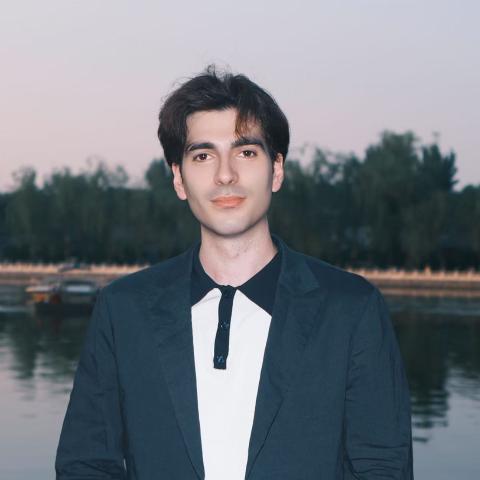 A man with dark hair wearing a dark blazer and a two-tone shirt stands in front of a body of water with trees in the background at dusk.