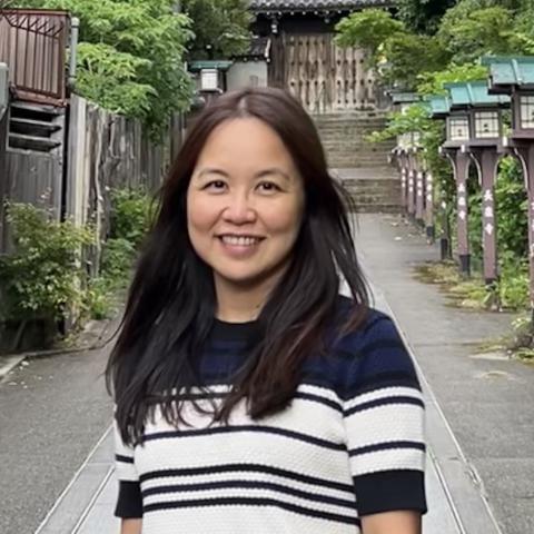 Woman with long dark hair stands smiling on a paved path lined with greenery and lanterns, with traditional wooden architecture in the background.