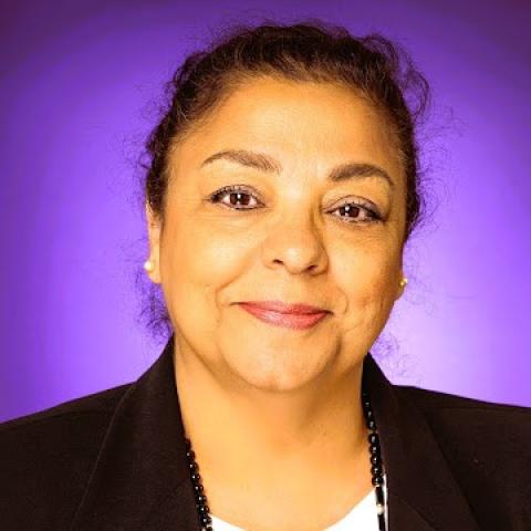 Portrait of a woman with dark hair pulled back, wearing a black blazer and a beaded necklace against a purple background.