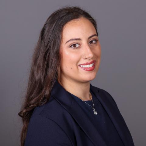 A smiling woman with long dark hair wearing a navy blue blazer and a pendant necklace.