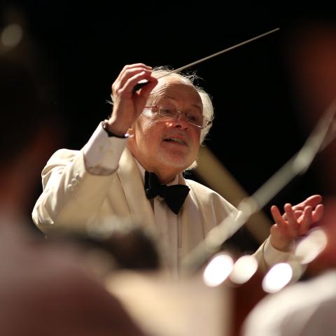 A conductor in a white suit and bow tie leads an orchestra, holding a baton and gesturing expressively with the other hand.