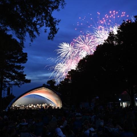 An outdoor concert takes place at dusk under a curved canopy, as fireworks light up the sky above trees and buildings in the background.