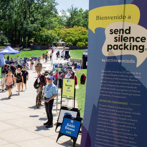 People walk through an outdoor exhibit called "Send Silence Packing" with informational signs and displays about mental health and suicide prevention.