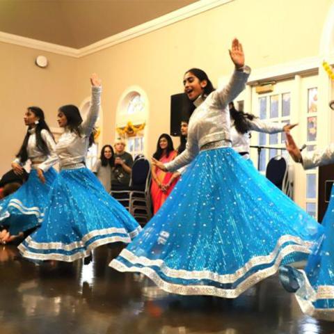 A group of women in blue and silver traditional attire perform a dance indoors, with onlookers watching in the background.