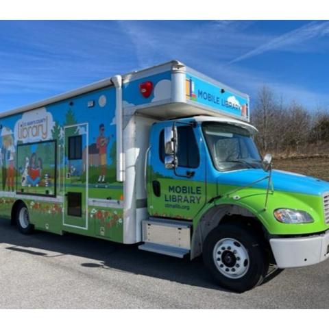 A colorful mobile library truck is parked on a paved road, featuring illustrations of books, people, and nature scenes on its side.