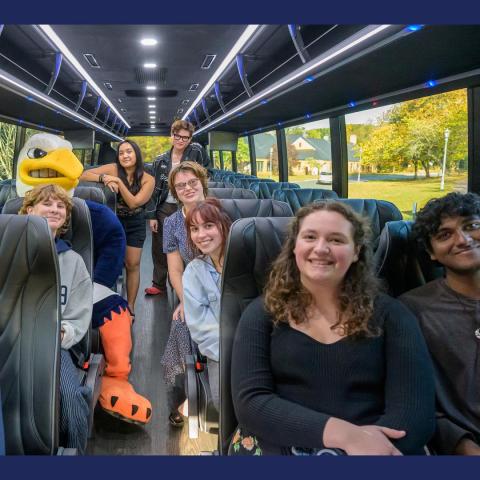 A group of smiling students and a person in an eagle mascot costume sit on a modern bus with blue interior lights and large windows showing trees outside.