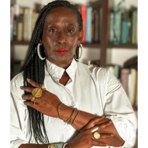 A woman with braided hair, wearing a white shirt and bold jewelry, poses with her arms crossed in front of a bookshelf.