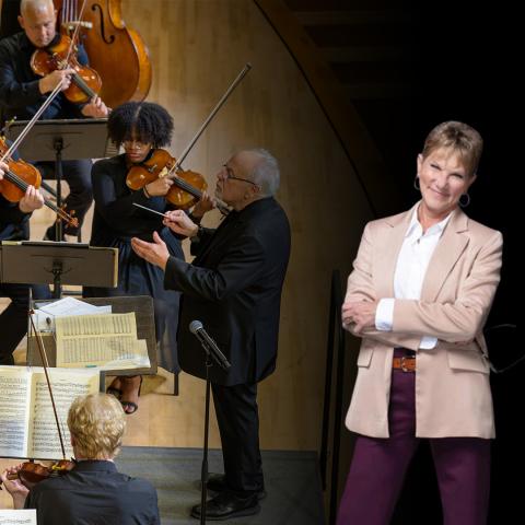 A conductor leads a string section during a performance, while a woman in business attire stands in the foreground, arms crossed and smiling.