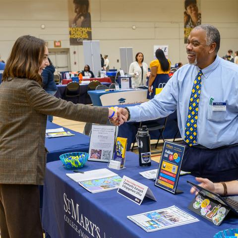 A woman and a man shake hands across a table at a college fair, with brochures, flyers, and informational materials displayed on the table.