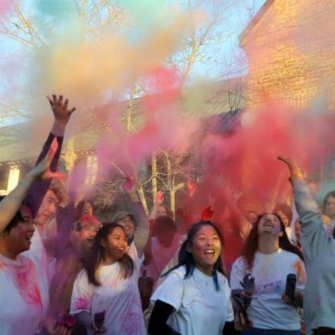 A group of people wearing white shirts throw colorful powder in the air outdoors, celebrating together in daylight.