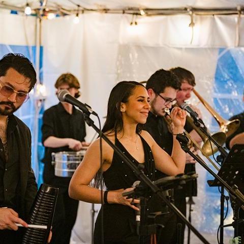 A band of six musicians, dressed in black, performs indoors with various instruments including brass, percussion, and microphones.