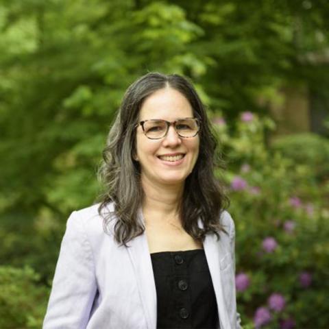 A woman with long brown hair and glasses, wearing a light-colored blazer over a black top, stands outdoors in front of greenery and flowering bushes.