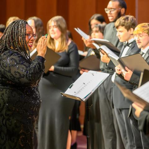 A choir director leads a group of singers dressed in formal black attire during a choral performance, with music stands and sheet music visible.