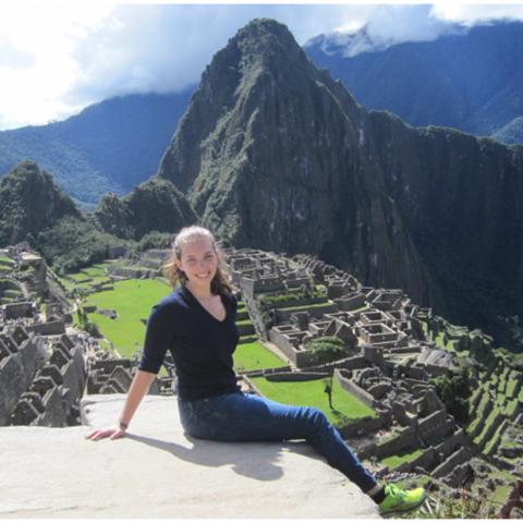 A person sits on a stone ledge overlooking the ancient ruins of Machu Picchu, with green terraces and a tall mountain in the background.
