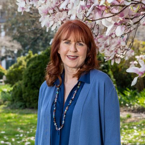 A woman with red hair wearing a blue outfit stands outdoors in front of blooming magnolia flowers and greenery.