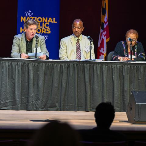 Three people sit at a long table on a stage during a panel discussion, with "The National Public Honors College" banner and a flag visible in the background.
