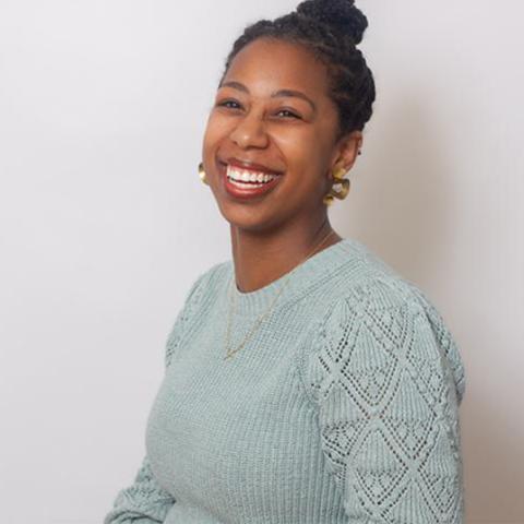 A woman with braided hair styled in a bun, wearing a light blue knit sweater, gold earrings, and a necklace, smiling against a plain light background.
