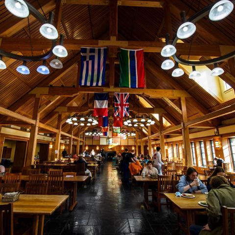 Large wooden dining hall with high ceilings, international flags hanging from beams, people sitting at tables eating, and bright overhead lights.