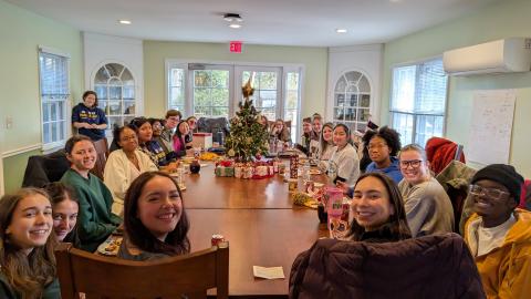 A group of people sit around a large table with food, drinks, and a small decorated Christmas tree in the center, gathered in a bright, cozy room.
