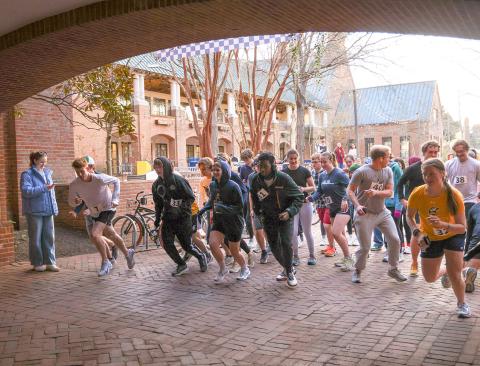 A group of runners at the starting line of a race in an outdoor courtyard, with a few spectators watching nearby.
