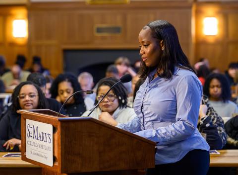 A woman stands at a podium labeled "St. Mary's College of Maryland" speaking to an audience seated in a wood-paneled room.