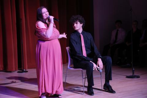 A woman in a pink dress sings into a microphone while standing next to a seated man in a black shirt and pink tie onstage, with red curtains and other people in the background.
