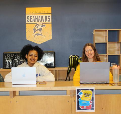 Two women sit at a table with laptops in front of a St. Mary’s College Seahawks sign; a "Brain Bee" event poster is visible on the front of the table.