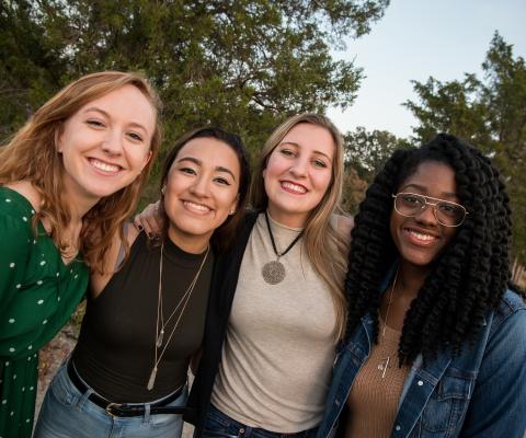 Four women stand close together outdoors, smiling at the camera, with trees and sky in the background.