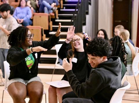 Four young adults sit in a circle indoors, smiling and giving high fives, with others seated in the background.