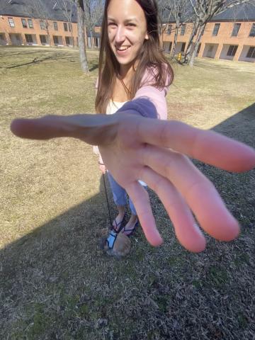 A woman stands outside on grass, reaching her hand toward the camera, with a small dog on a leash near her feet. Brick buildings and leafless trees are in the background.