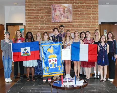 A group of people stands indoors, holding Haitian, French, and society flags, with documents and candles visible on a table in front of them.