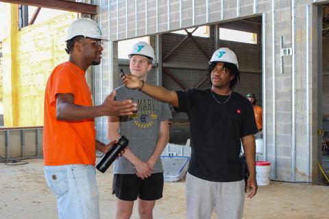 Three people wearing hard hats stand inside a building under construction; one gestures while another holds a recorder toward him.