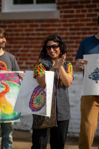 A woman stands outdoors, smiling and clapping, holding colorful artwork. She is flanked by two people also holding art, with a brick wall in the background.
