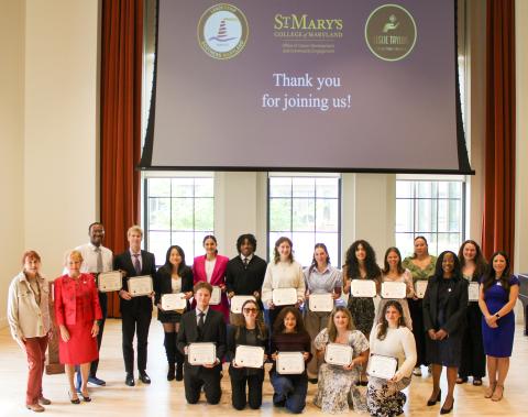 A group of people pose indoors, holding certificates, in front of a screen that displays “Thank you for joining us!” and logos for St. Mary’s College of Maryland and Issue TWIQI.
