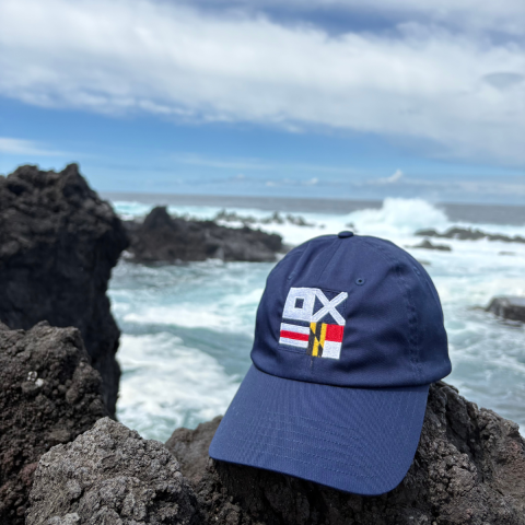 A blue baseball cap with multicolored flag designs rests on dark volcanic rocks by the ocean, with waves and a cloudy sky in the background.