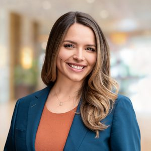 Smiling woman with long brown hair, wearing a blue blazer and orange top, stands in a softly lit office environment, conveying a professional and warm tone.