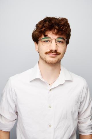 A young man with curly brown hair, glasses, and a mustache wearing a white button-up shirt stands against a plain light gray background.
