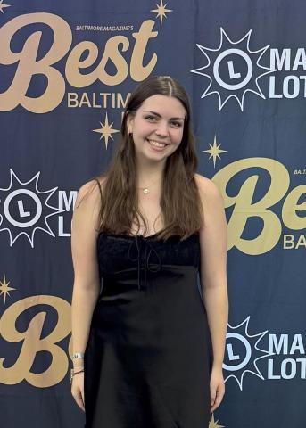 A woman in a black dress stands smiling in front of a backdrop that reads "Best of Baltimore" and "Maryland Lottery.