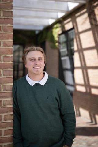 A young man with light brown hair, wearing a green sweater over a white collared shirt, stands and smiles while leaning against a brick wall in an outdoor setting.