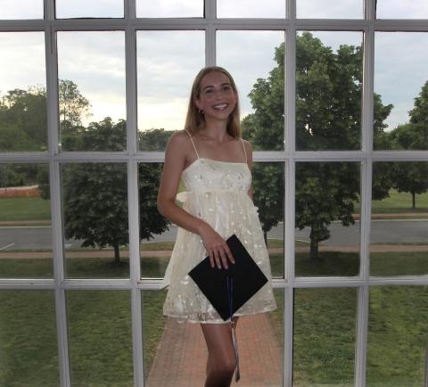 A young woman in a white dress stands in front of large windowpanes, holding a graduation cap, with trees and grass visible outside.