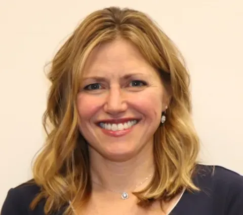 A woman with shoulder-length blonde hair smiles at the camera against a plain, light-colored background. She is wearing a dark top and a necklace.
