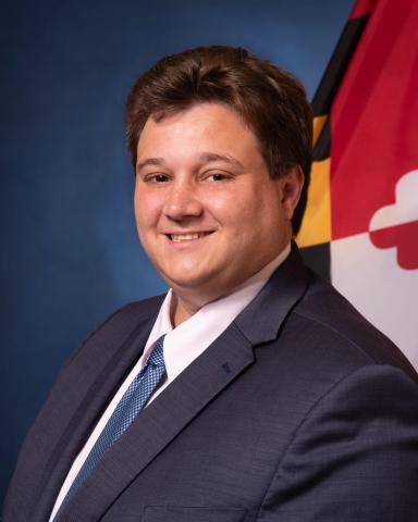 Man in a suit and tie poses in front of a blue background with part of a colorful flag visible on the right.
