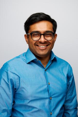 A man with short dark hair, wearing glasses and a blue button-up shirt, smiles at the camera against a plain light background.