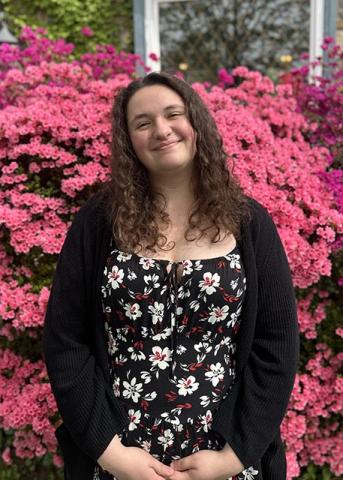 A person with curly brown hair stands in front of a large, vibrant pink flowering bush, wearing a black floral dress and a black cardigan, smiling at the camera.