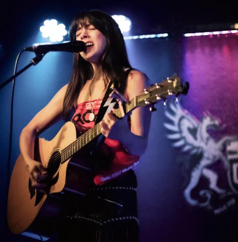 A woman sings while playing an acoustic guitar on stage, with a microphone in front of her and stage lights in the background.