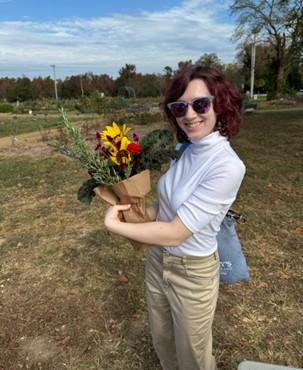 A person wearing sunglasses and a white turtleneck holds a bouquet of flowers outdoors on a sunny day.