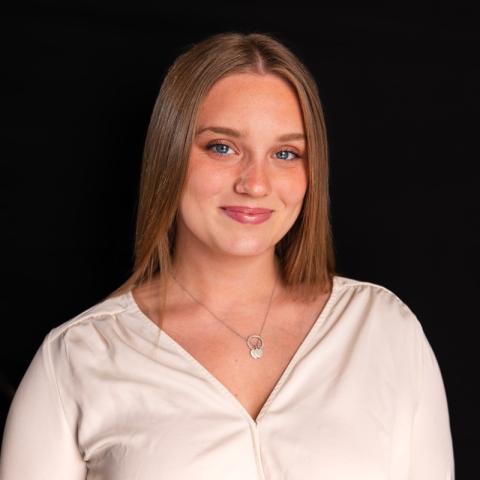 A woman with straight, light brown hair and a light blouse smiles slightly, standing against a solid black background.