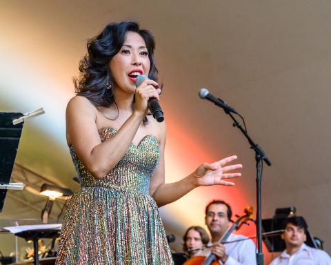 A woman in a sparkly dress sings into a microphone on stage, with musicians and music stands visible in the background.
