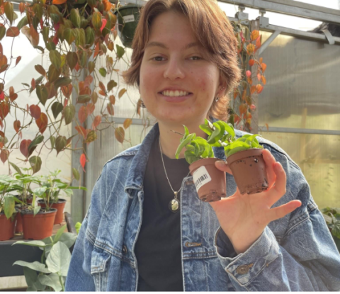 A person in a denim jacket smiles while holding two small potted plants in a greenhouse. The background features hanging and potted plants, creating a lush, vibrant atmosphere.