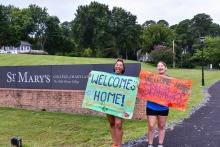 Two smcm Orientation Leaders hold signs that read "Welcome Home" and "Welcome Home Seahawks" in front of the College Drive on new student move-in day. 